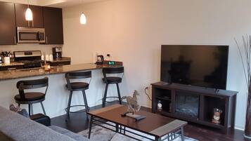 Een moderne keuken met donkere kastjes, een roestvrijstalen koelkast, een magnetron en een koffiezetapparaat. Er is een zithoek met een bank en een tafel, en een flatscreen televisie op een standaard.
