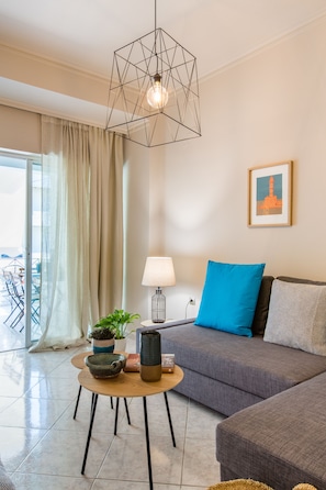 Una sala de estar moderna con un sofá gris, una almohada azul, una lámpara colgante geométrica y una pequeña mesa redonda con un cuenco y una planta.