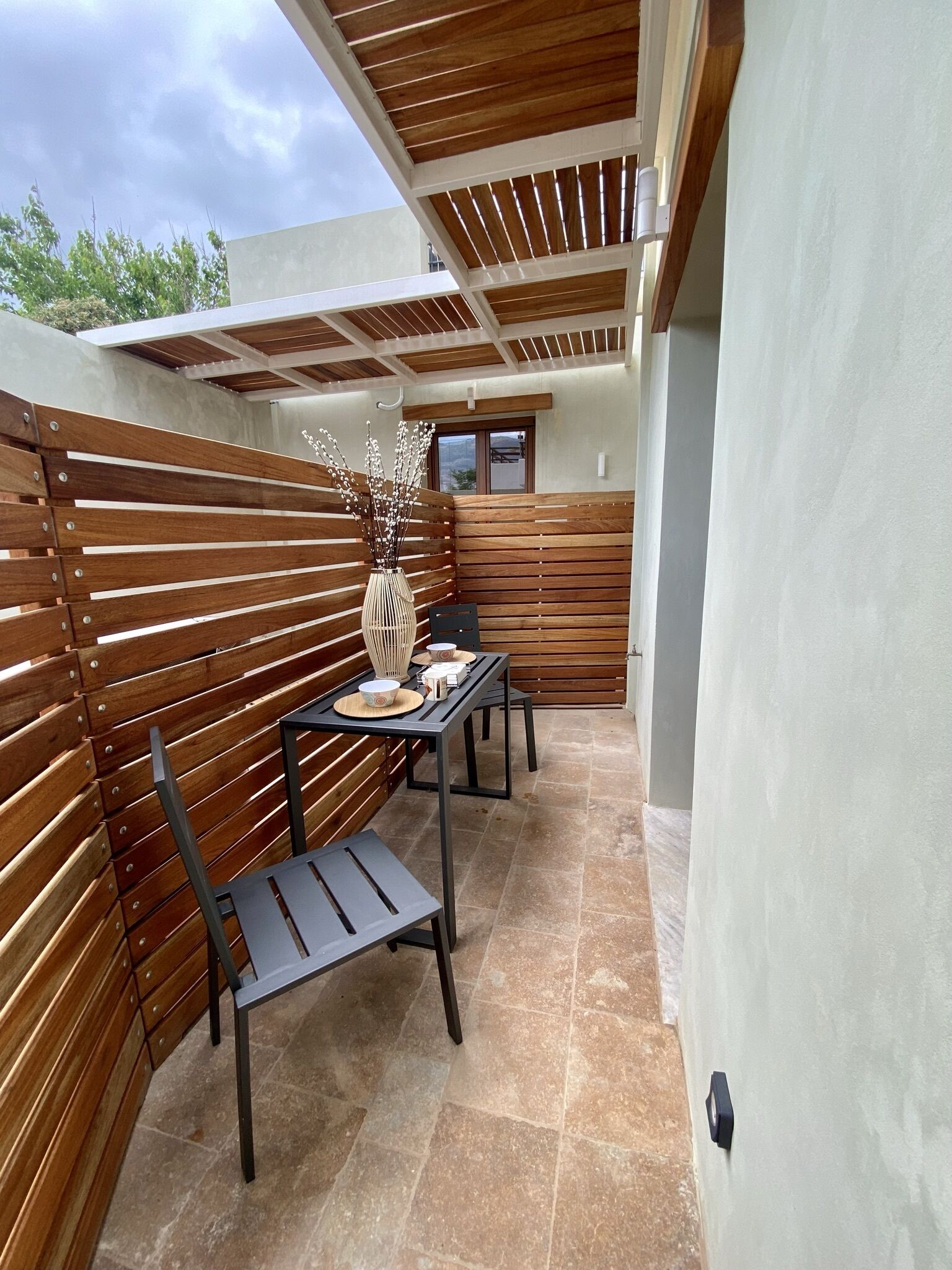 An outdoor patio with a wooden slatted wall, a table with chairs, and a vase with branches.