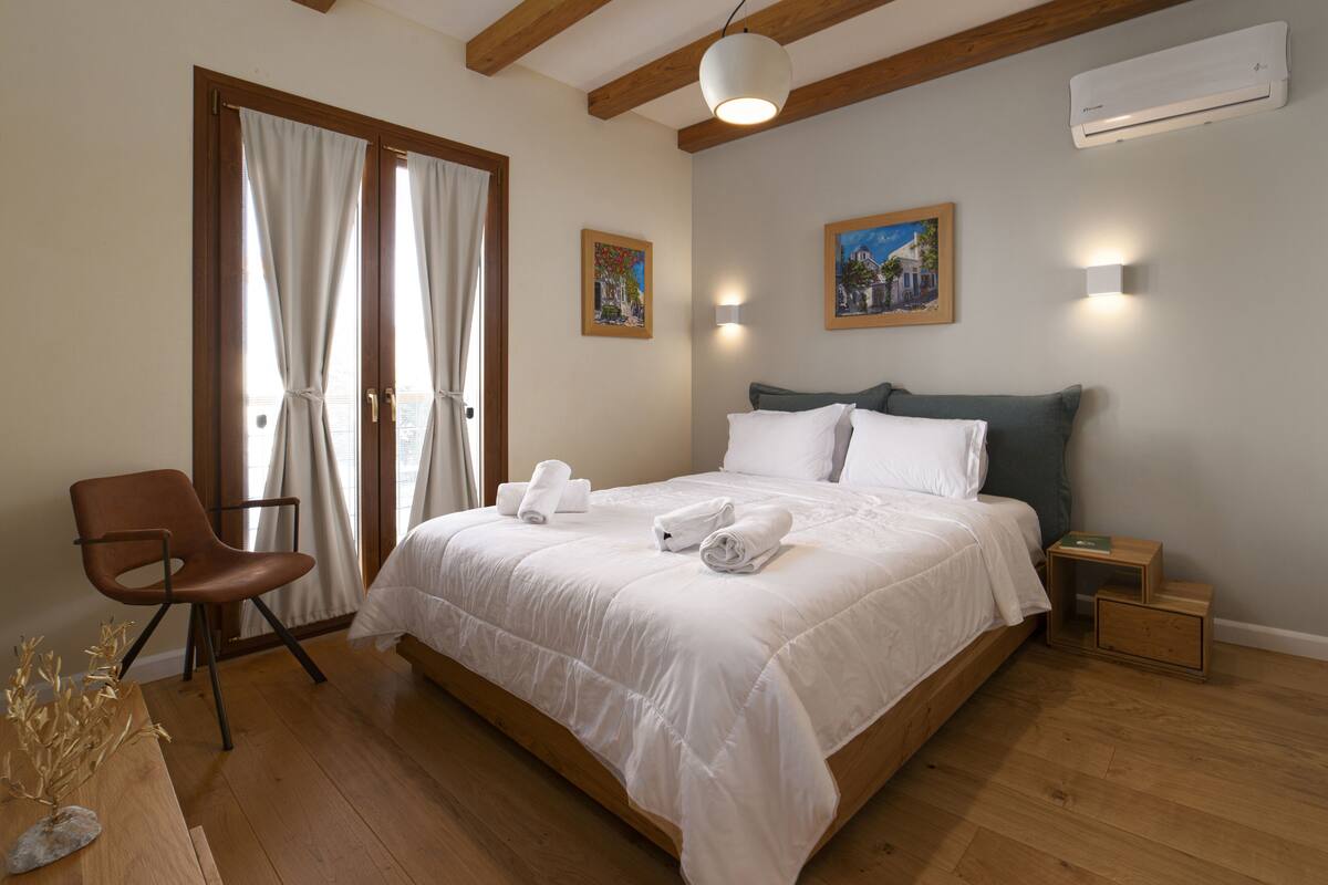 A bedroom with a wooden bed, white bedding, a wooden chair, and a small table with a vase.