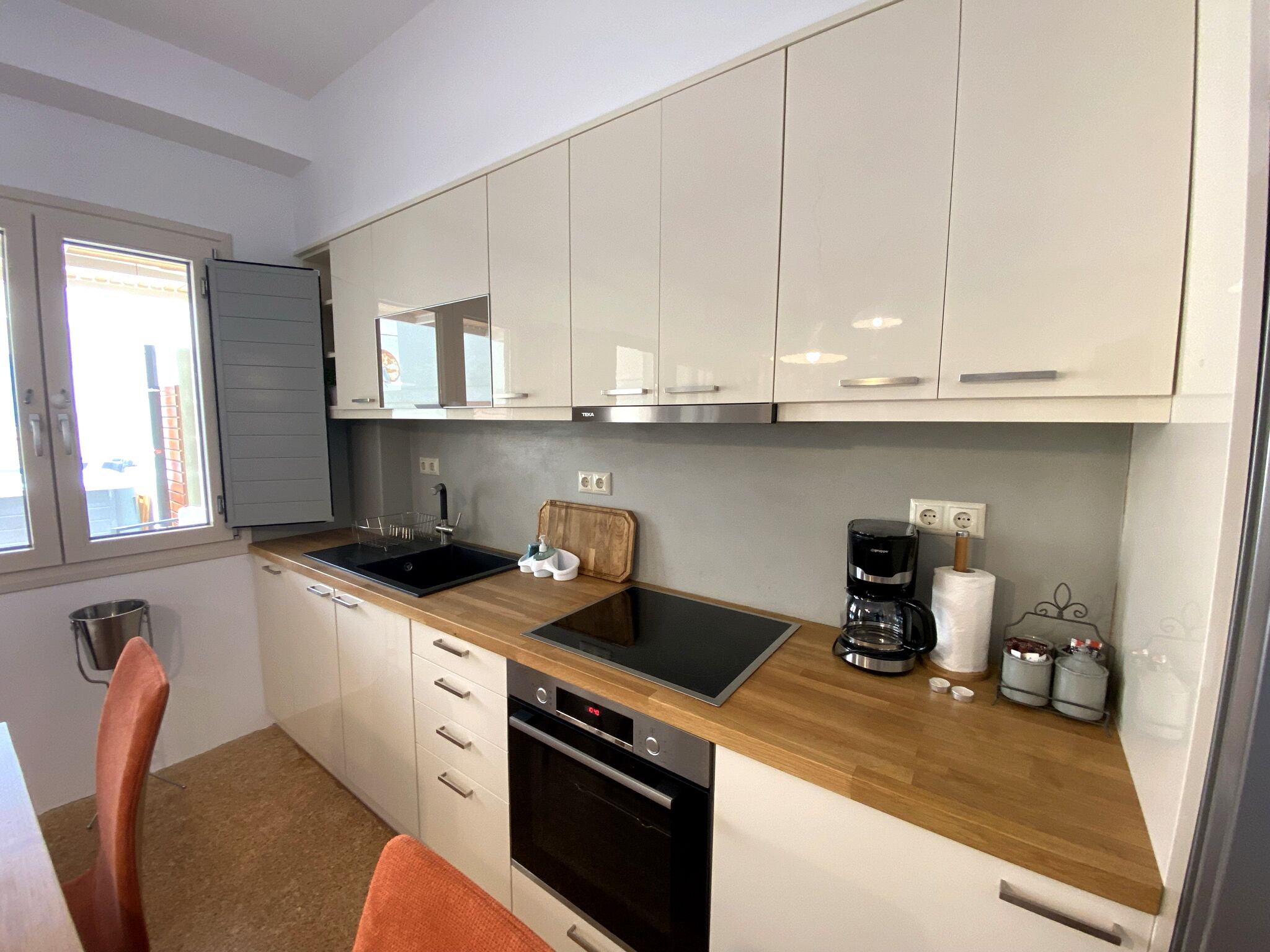 Cocina moderna con armarios blancos, un lavamanos negro, encimera de madera, cafetera y una ventana con persianas.