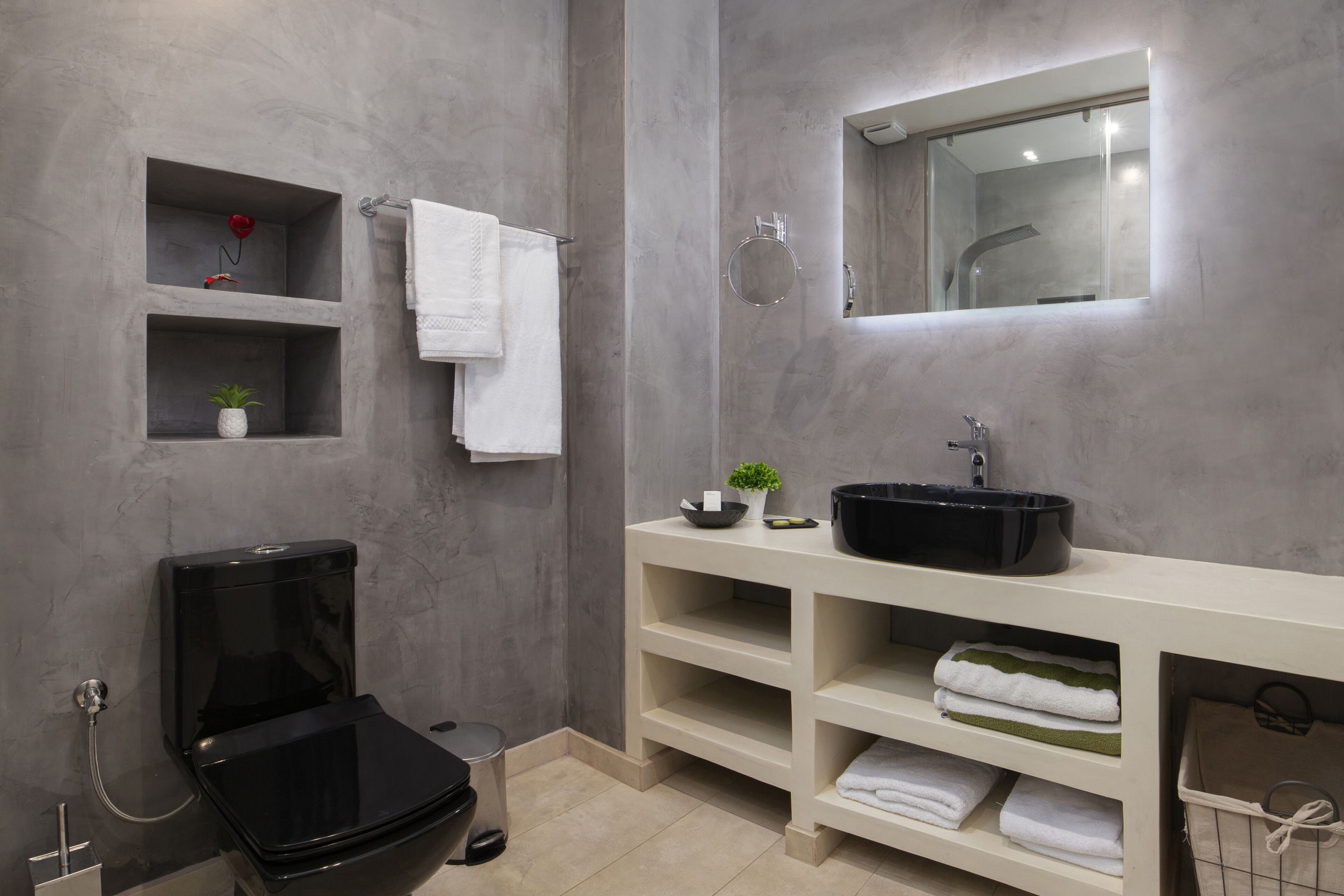 Ein modernes Badezimmer mit einer schwarzen Toilette, einem weißen Waschbecken mit schwarzer Schüssel und einem Spiegel darüber. Es gibt Regale mit Handtüchern und einer kleinen Pflanze.