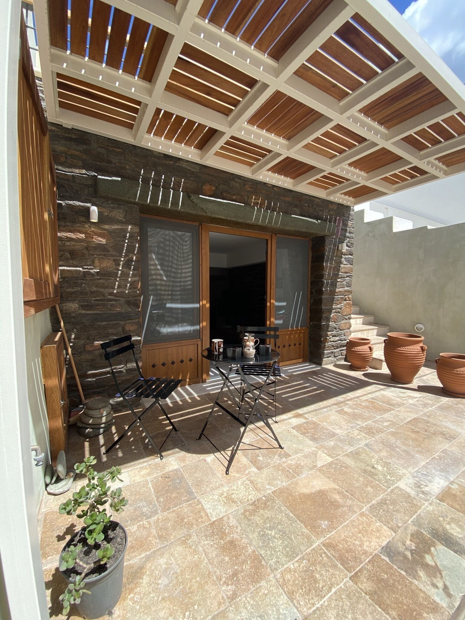 A patio with a stone wall, a wooden pergola, a table with chairs, and terracotta pots.