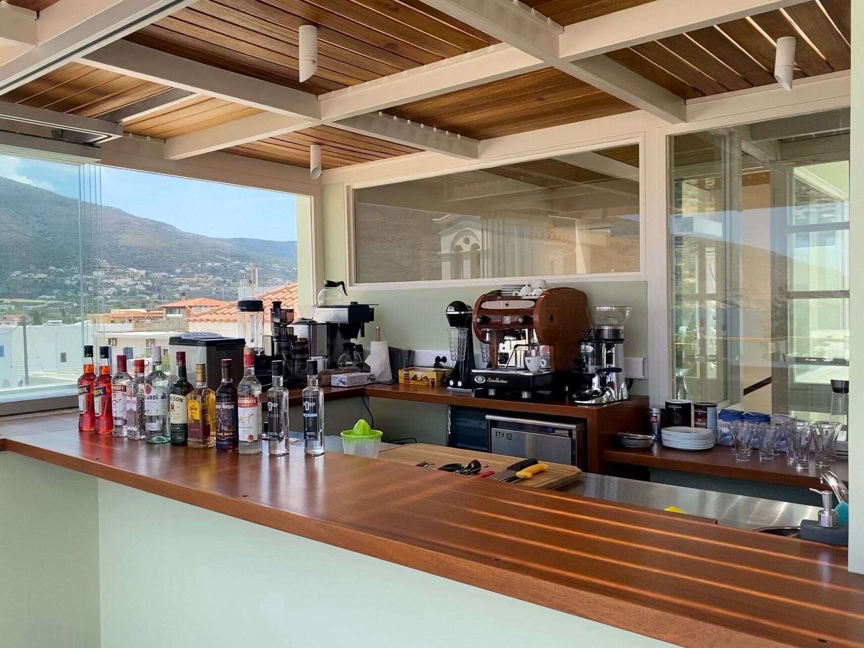 A bar area with a coffee machine, various bottles, and a view of a mountainous landscape through large windows.