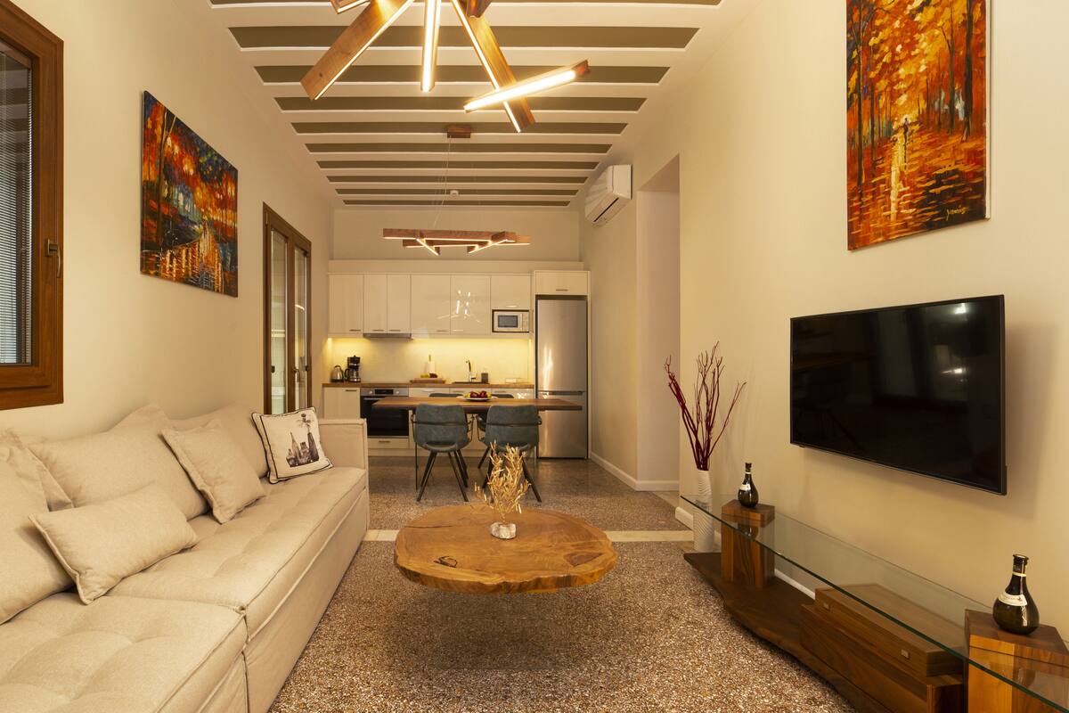 A modern living room with a sofa, coffee table, and TV. There is a kitchen in the background with a fridge and microwave.
