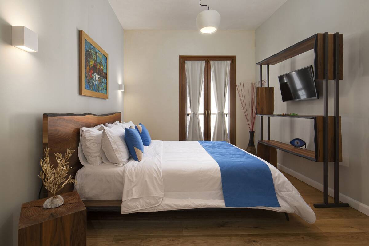 A bedroom with a bed, a TV mounted on a wooden shelf, and a painting on the wall.