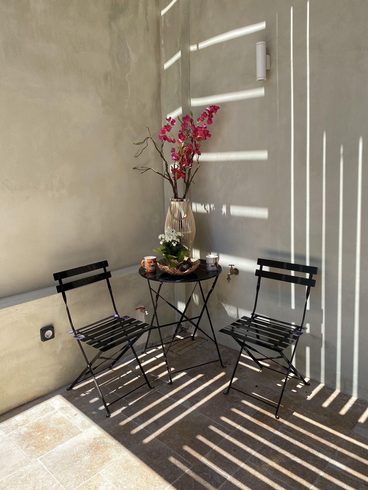 A small outdoor seating area with two metal chairs and a table, a vase with pink flowers, and a wall-mounted light fixture.