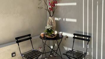 Un pequeño espacio al aire libre con dos sillas de metal y una mesa, un florero con flores rosas, y una lámpara fijada a la pared.