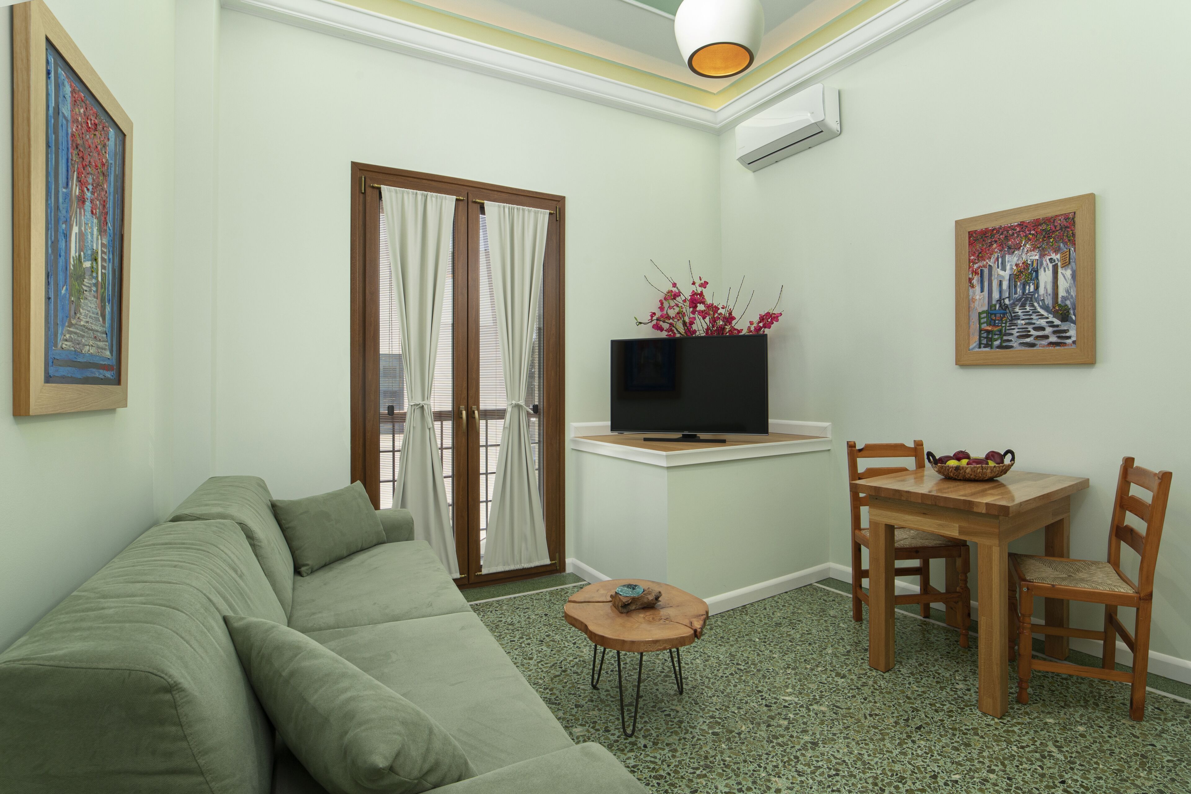 A living room with a green sofa, a wooden dining table with chairs, a small round table with a bowl, and a flat-screen TV on a stand.