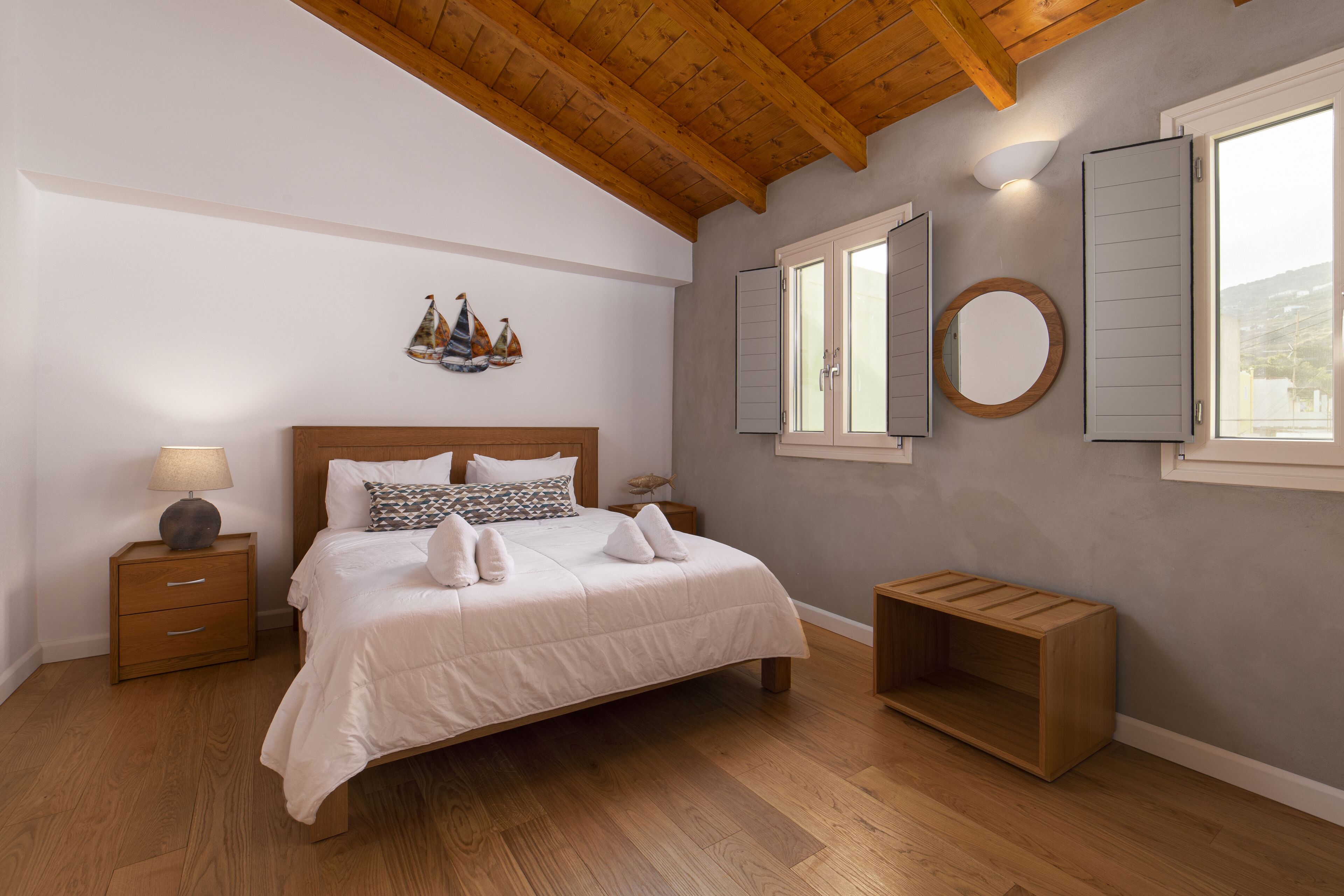 Un dormitorio con techo de madera, una cama con ropa blanca, una mesita de noche con lámpara, un banco de madera y un espejo montado en la pared.