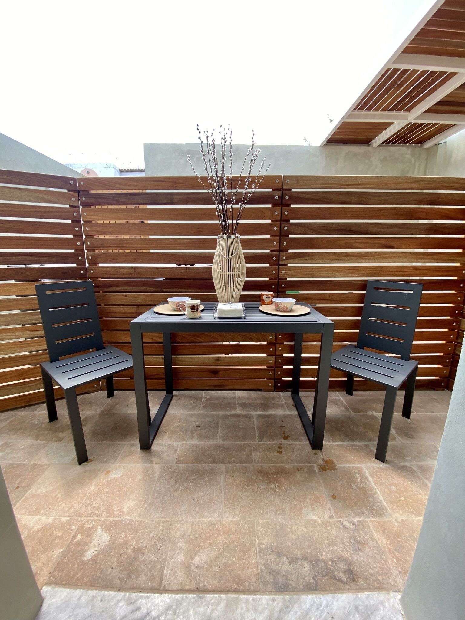 An outdoor patio area with a table and two chairs, a vase with branches, and a wooden slatted wall.