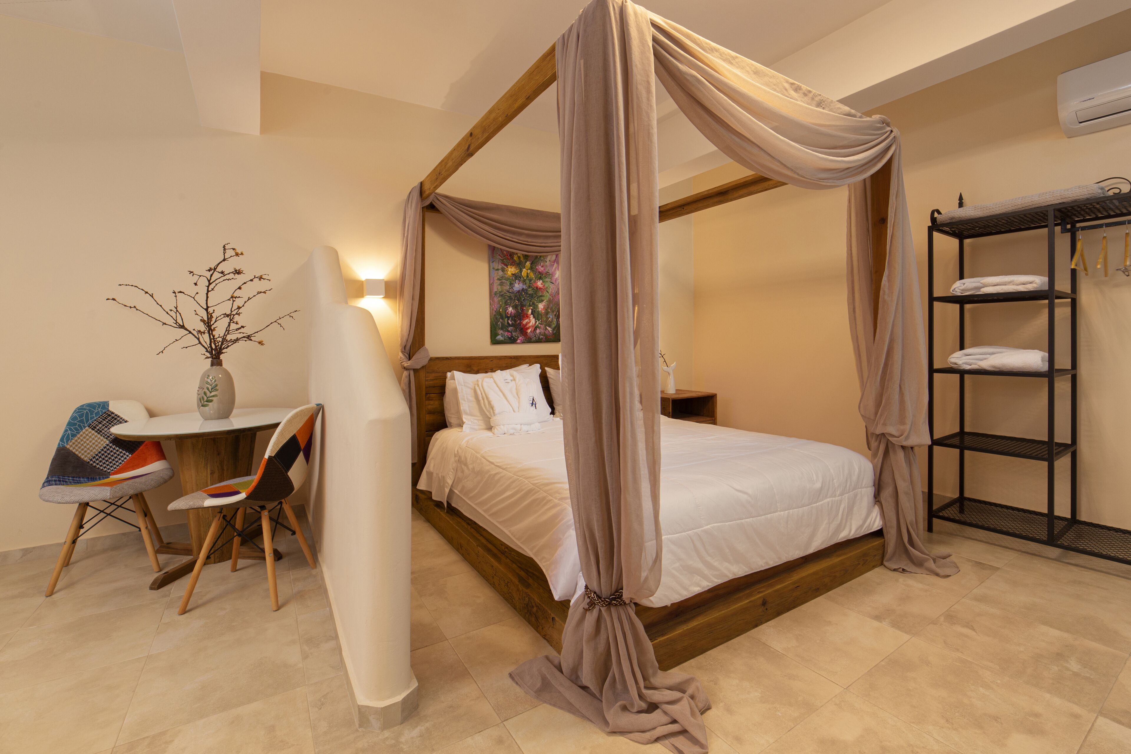 Una cama con dosel sobre cuatro columnas, con estructura de madera, una mesita de noche con un florero, una silla y un estante con toallas.