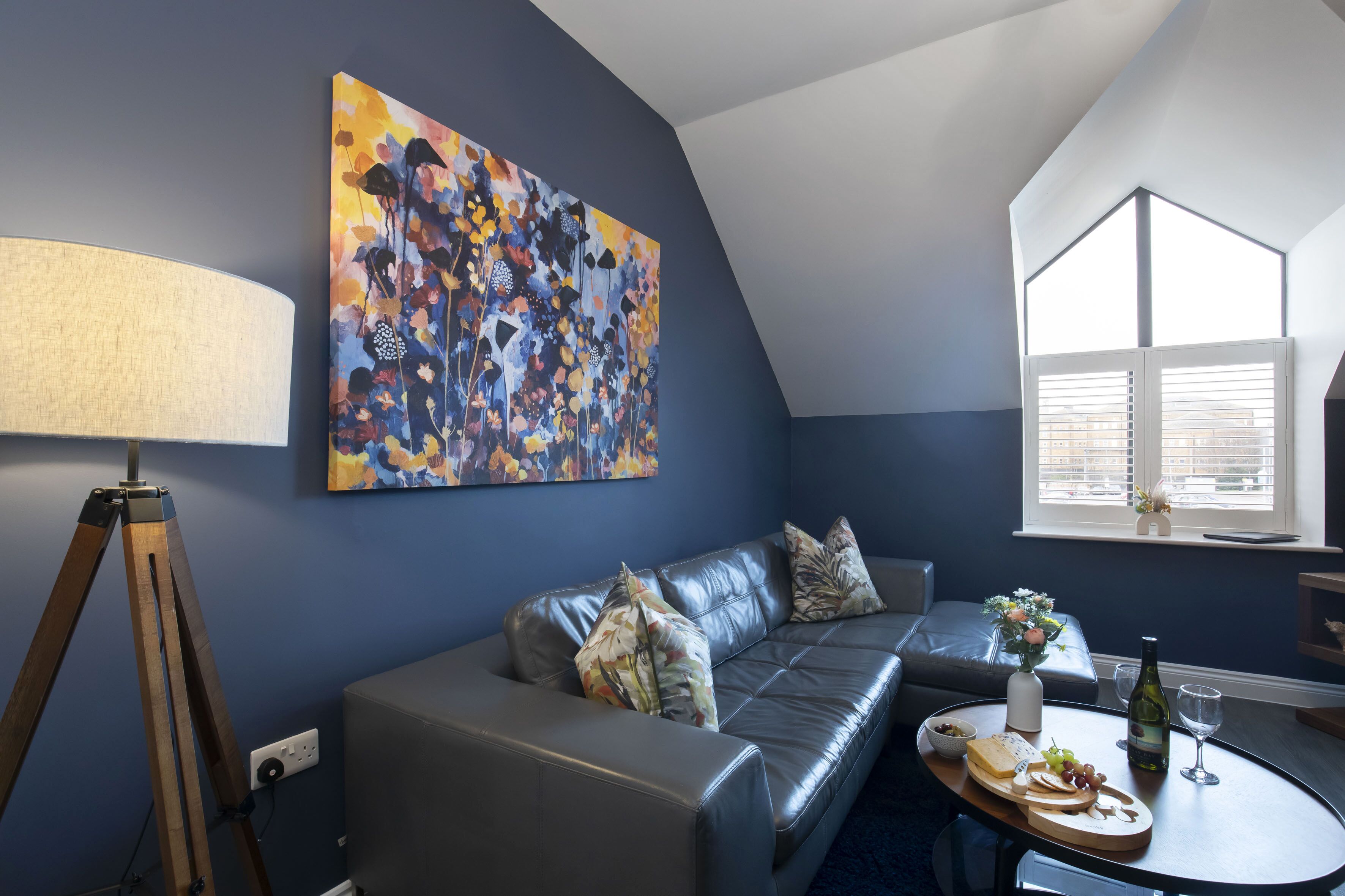 Een moderne woonkamer met een donkere blauwe muur, een groot abstract schilderij, een leren bank, een ronde salontafel met eten en drinken, en een driepoot lamp.