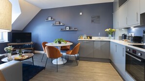 A modern kitchen with grey cabinets, a dining area with a round table and orange chairs, and a TV in the background. - Elliot Oliver - Loft Style 2 Bedroom Apartment With Parking In The Docks (Gloucester)