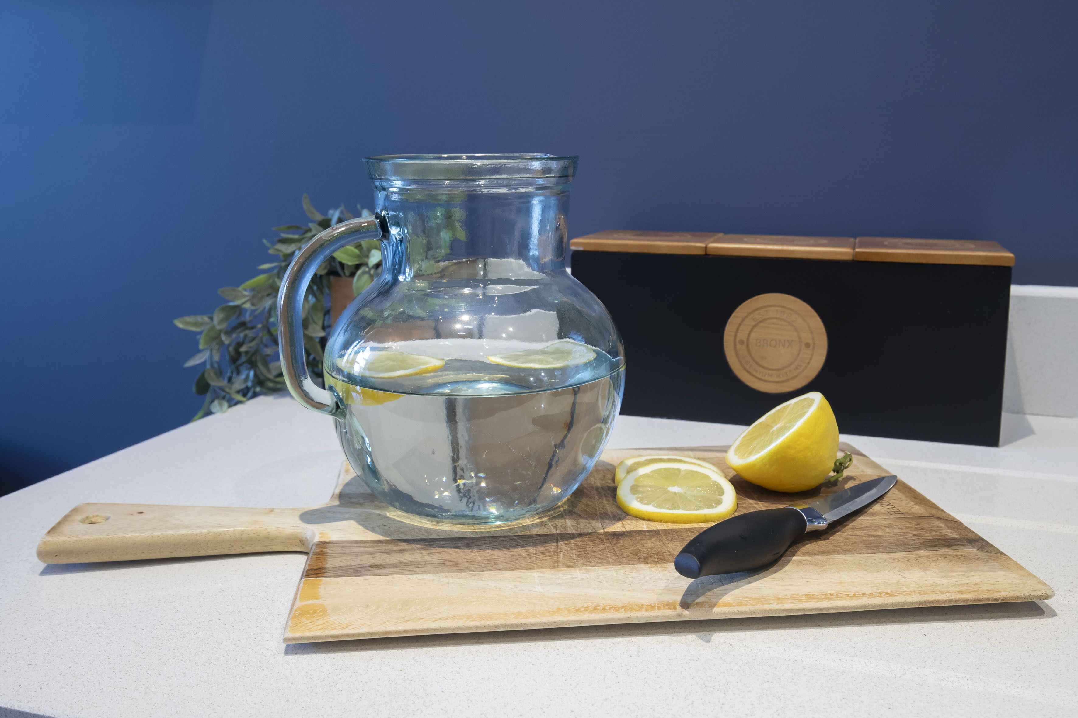 A glass pitcher with water and lemon slices on a wooden cutting board, a knife, and a box with a circular emblem.