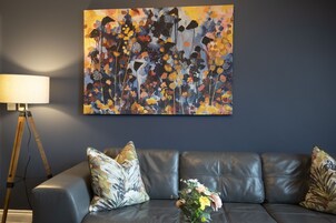 Una sala de estar moderna con un sofá de cuero oscuro, cojines estampados, una lámpara de pie y un cuadro abstracto grande en la pared.