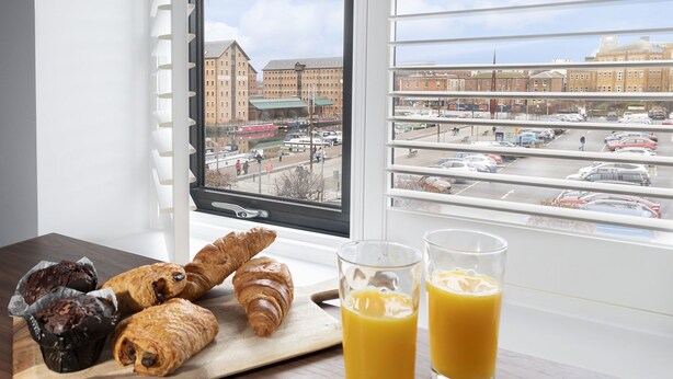 A breakfast setup with croissants and orange juice by a window overlooking a parking lot and buildings. - Elliot Oliver - Loft Style 2 Bedroom Apartment With Parking In The Docks (Gloucester)
