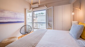 Una camera da letto con un letto, una sedia, un poggiapiedi in vimini, un quadro e vista sull'esterno.