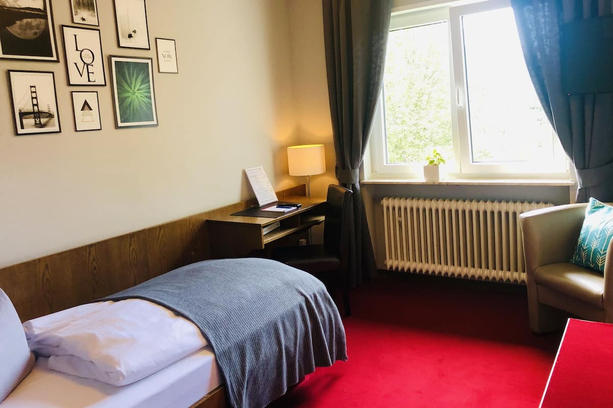 A hotel room with a bed, a desk with a lamp, a window with curtains, a radiator, and a view of greenery outside.