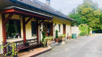 Ein gelbes Gebäude mit rotem Dach und einer Veranda mit Bänken. Auf der Veranda stehen Pflanzgefäße und davor eine Straße.