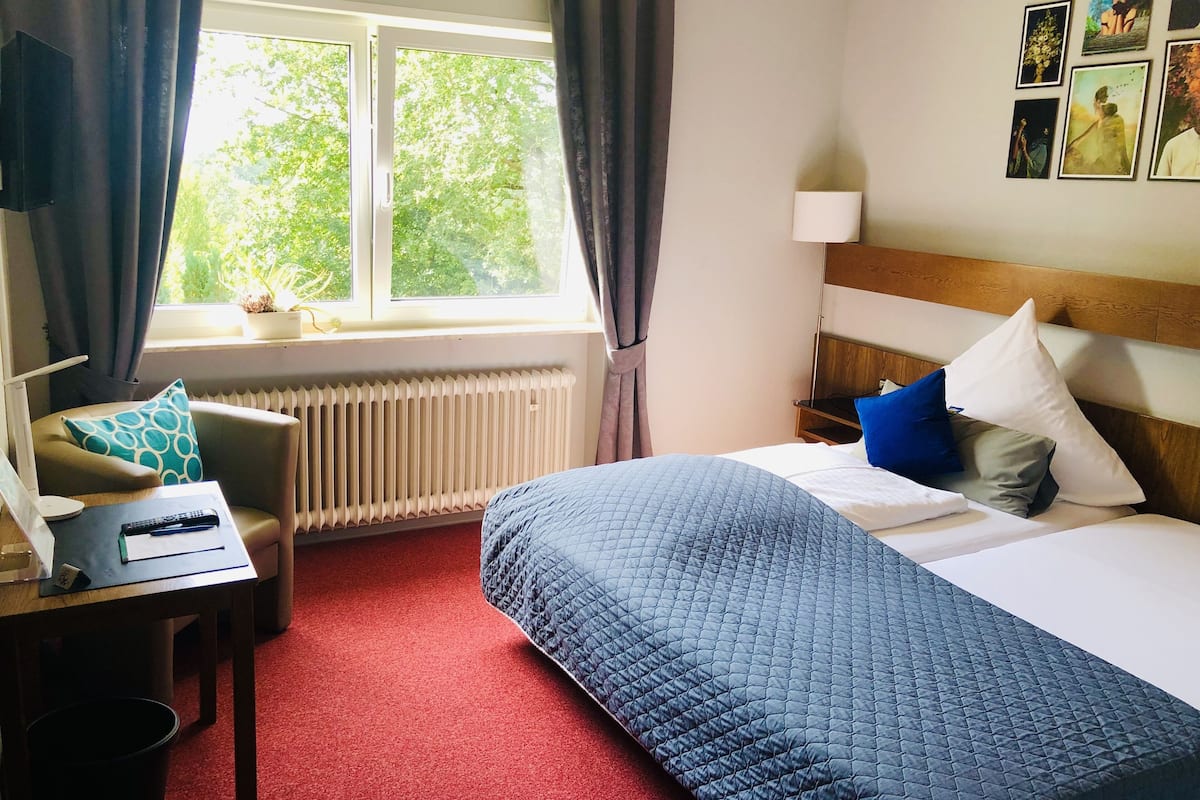 A hotel room with a bed, a desk with a lamp, a chair, a window with curtains, and a radiator.