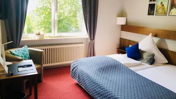 A hotel room with a bed, a desk with a lamp, a chair, a window with curtains, and a radiator.