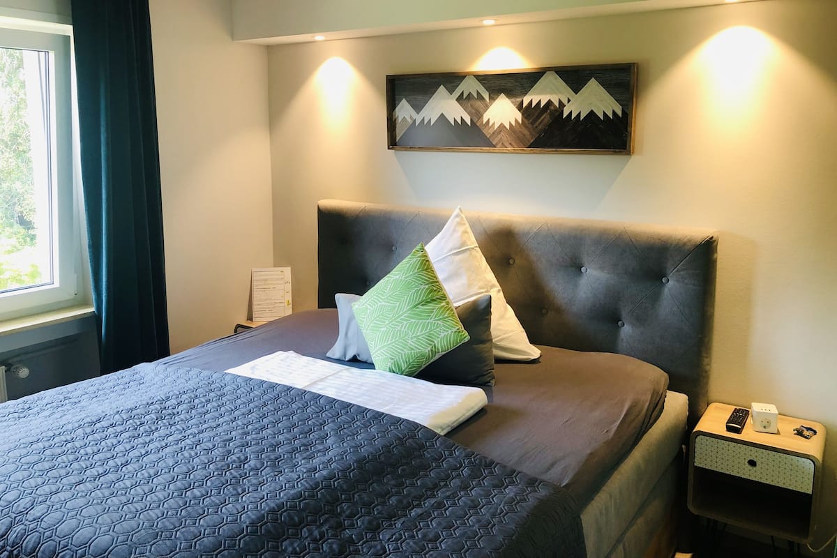 A neatly made bed with a dark quilt, a green pillow, and a white pillow. There is a wall-mounted painting of mountains and a bedside table with a remote control and a small box.