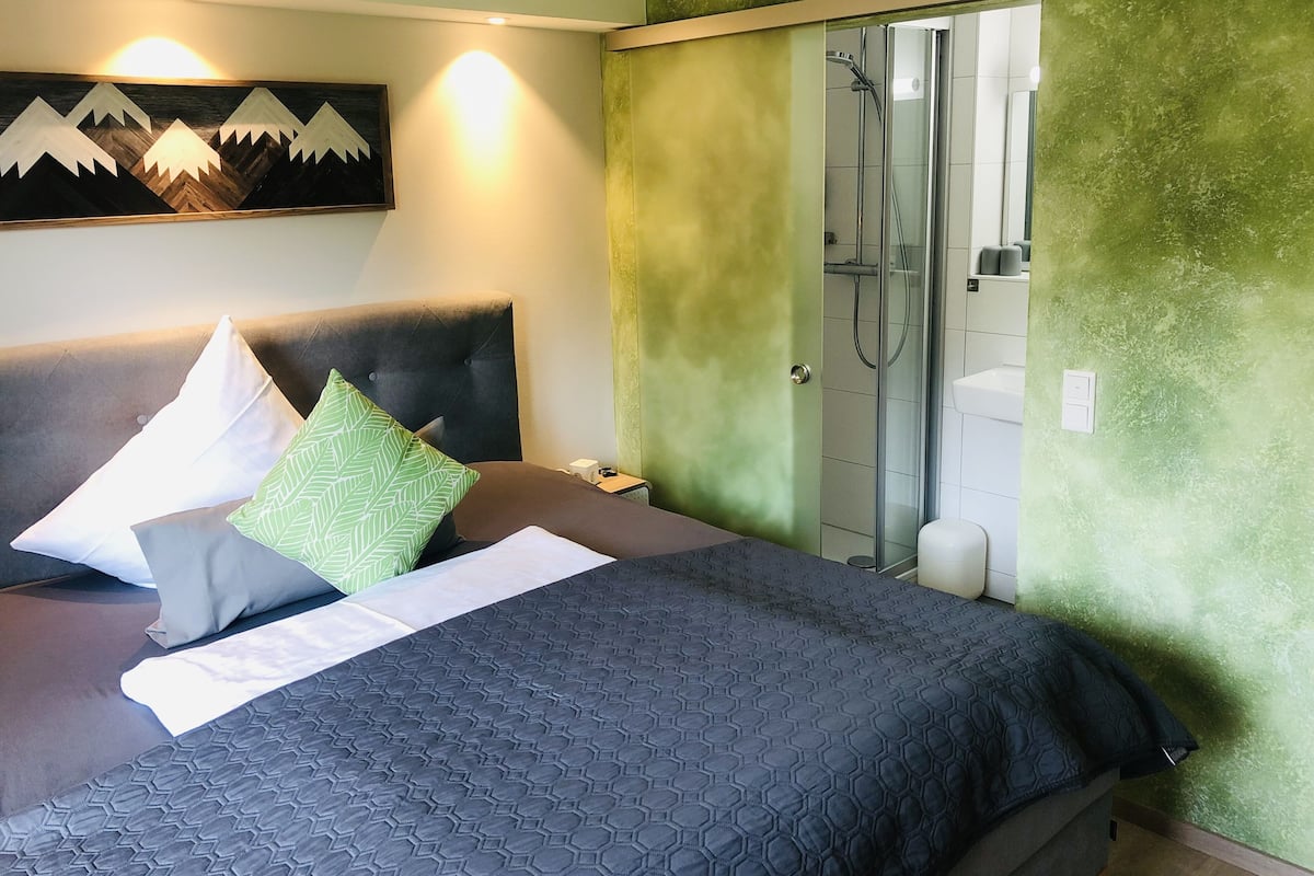 A hotel room with a bed, a nightstand, a green textured wall, a shower area, and a painting of mountains.