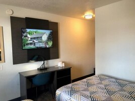 Habitación de hotel con cama, escritorio, silla, televisor montado en la pared y lámpara de pared.