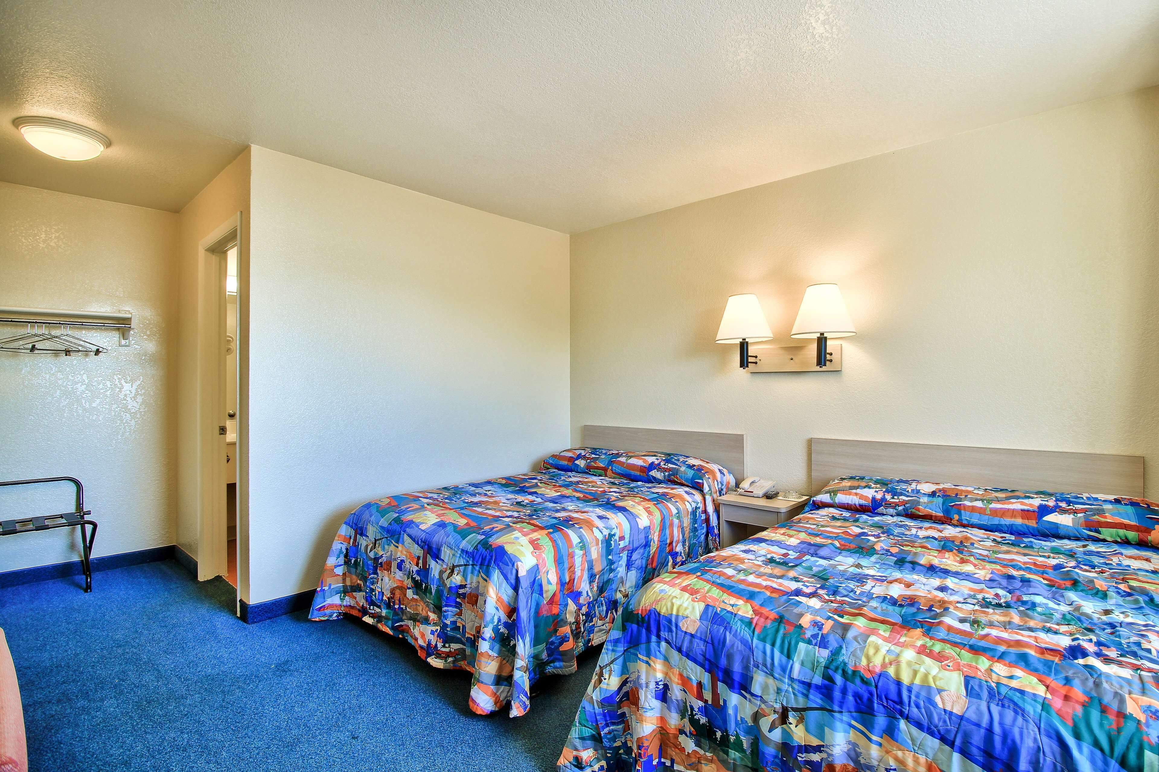 A hotel room with two beds, a nightstand, a lamp, and a coat rack.