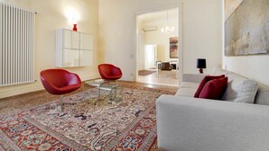 A living room with a sofa, two red chairs, a patterned rug, a white cabinet, and a dining area in the background.