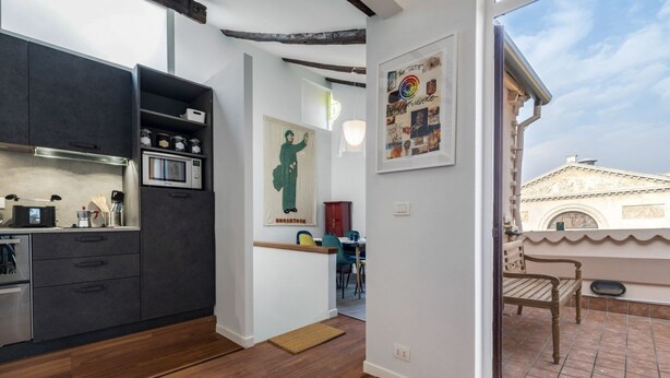 A modern kitchen with dark cabinets, a microwave, and a wooden floor. There is a dining area with a table and chairs, and a balcony with a view of a building. - CA' SVEVA VENICE APARTMENT M0270429660 (Venezia)