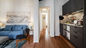 A modern living room with a blue sofa, a wooden chair, and a black ottoman. There is a painting on the wall and a kitchen with dark cabinets and stainless steel appliances. - CA' SVEVA VENICE APARTMENT M0270429660 (Venezia)