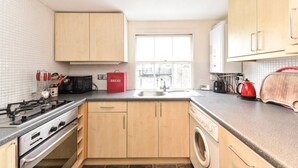 A compact kitchen with wooden cabinets, a gas stove, a microwave, a washing machine, and a window providing natural light. - Smart flat near Pimlico Tube (London)