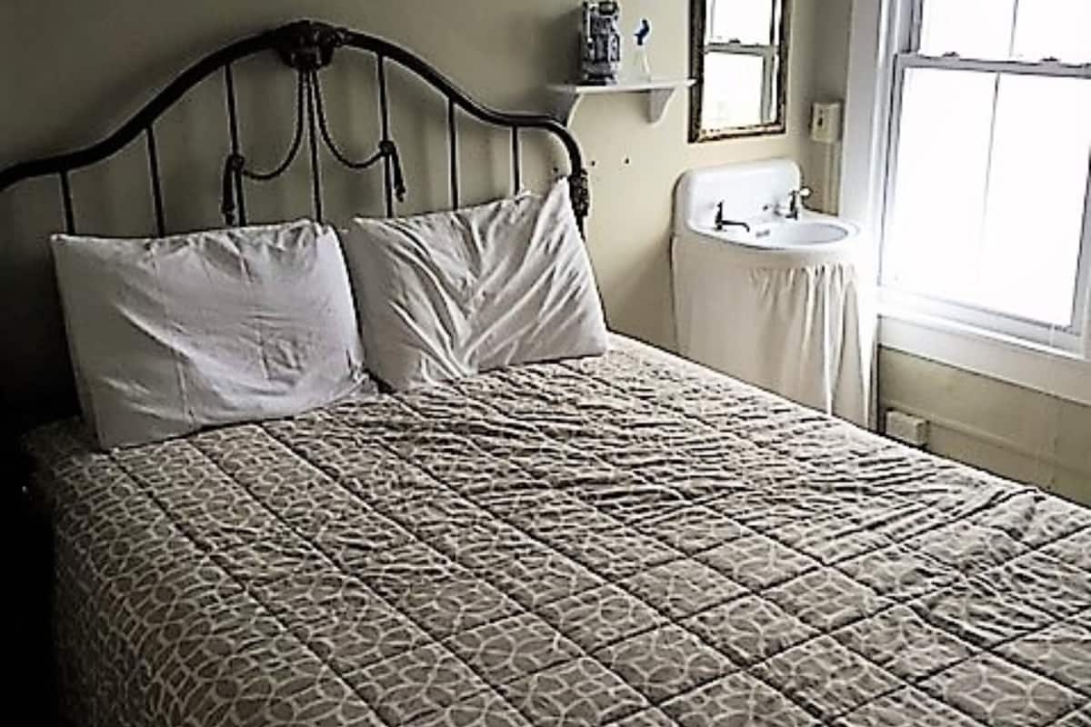 Una cama con una colcha estampada, una mesita de noche, un espejo adosado a la pared y una ventana con cortinas.
