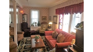 A living room with a red sofa, two armchairs, a coffee table with a plant, and a lamp. There is a window with curtains and a door with an exit sign. - Bath Avenue House (Ocean Grove)