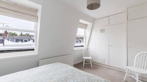 Een slaapkamer met een bed, twee witte stoelen, een kledingkast en een raam met uitzicht op gebouwen.