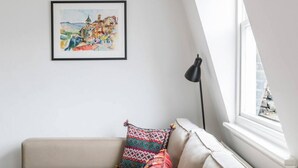 Een gezellige kamer met een beige bank, kleurrijke kussens, een lijstwerk met een schilderij en een zwarte vloerlamp.