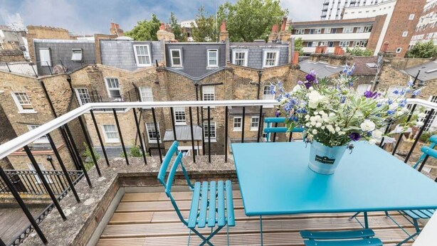 Een terras met een blauwe tafel en bijpassende stoelen, een bloevas en uitzicht op terrashuizen.