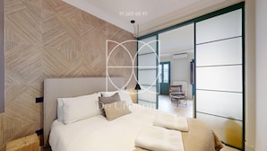 Ein modernes Schlafzimmer mit einer Wand im geometrischen Muster, einem Bett mit weißer Bettwäsche und Blick auf eine Stadtlandschaft durch eine Schiebetür.