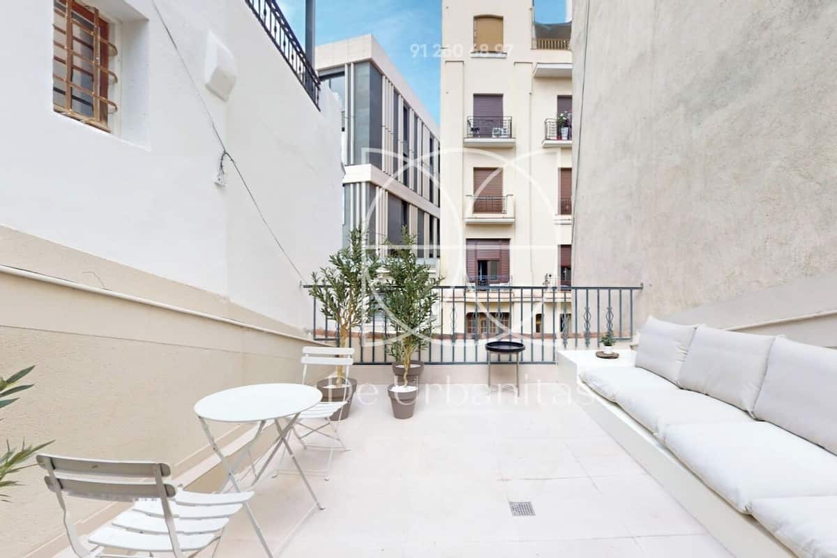 A narrow courtyard with a white sofa, a small round table, and two chairs. There are potted plants and a balcony railing visible.