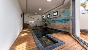 A modern indoor swimming pool with a wooden deck, a mural of a beach scene, and a view of the sky through a large window. - ARES - Vue Mer - Accès direct plage (Agde)
