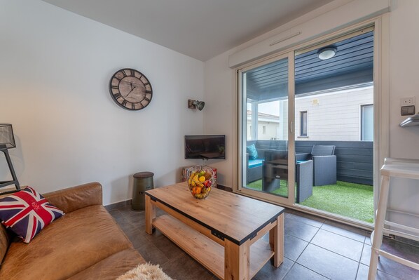 A living room with a wooden table, a sofa with a Union Jack pillow, a wall clock, and a TV.