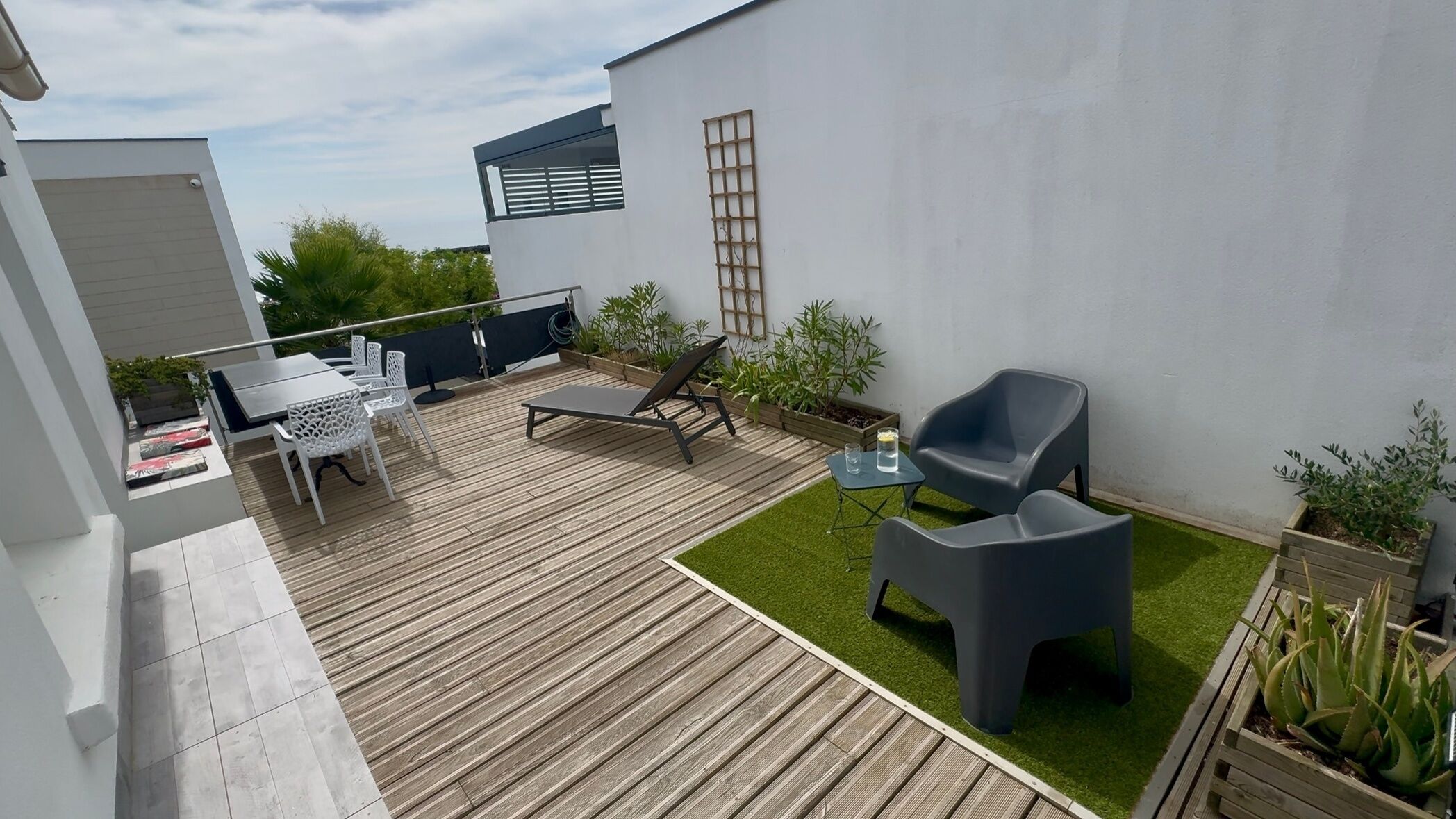 Eine moderne Terrassenanlage im Freien mit Holzboden, einem weißen Tisch mit Stühlen, einer Liege und einem kleinen Tisch mit einem Glas.