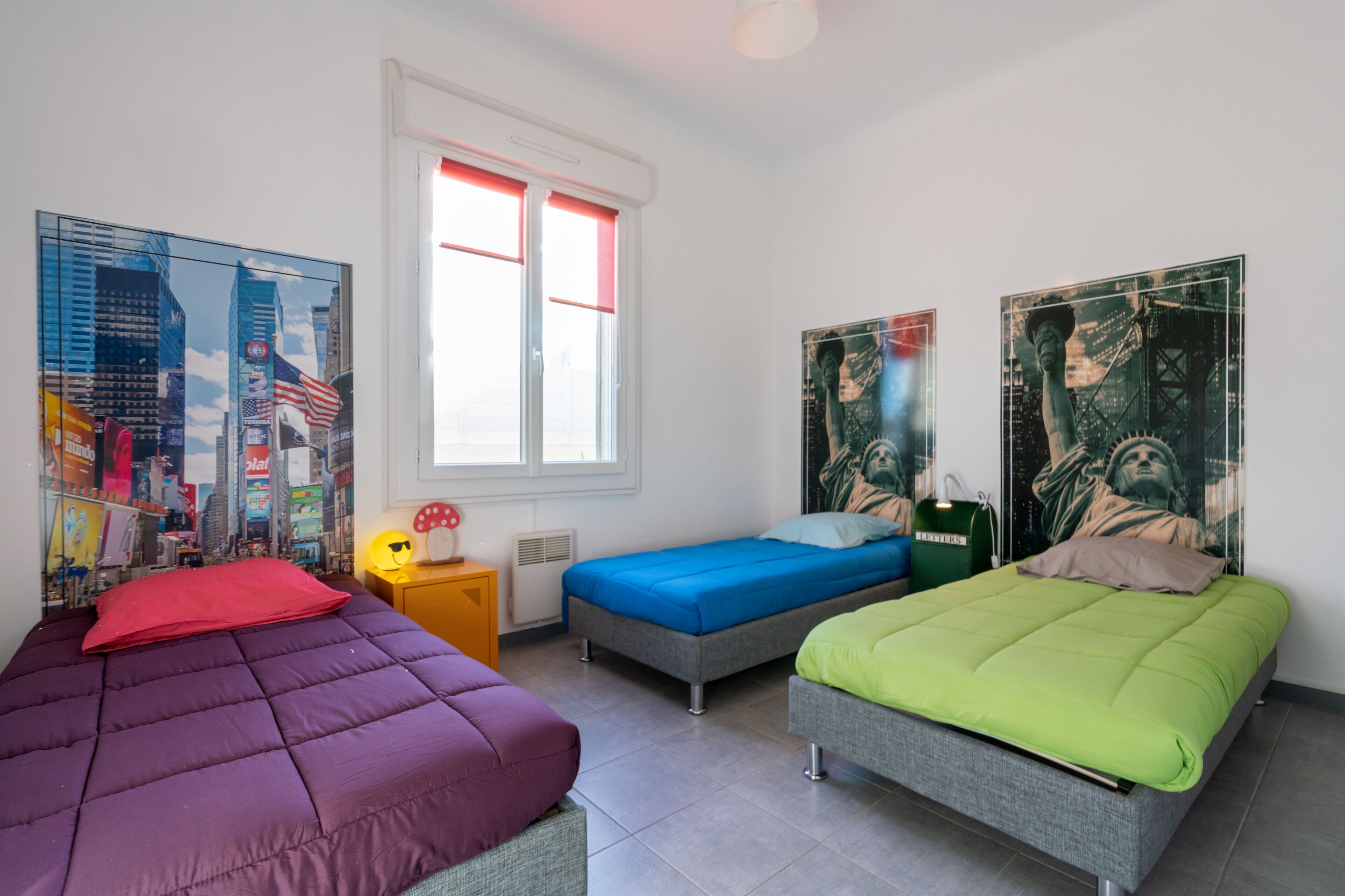 A room with three beds, each with a different colored bedspread. There are posters on the walls, a window with red blinds, and a small table with a lamp.
