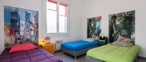A room with three beds, each with a different colored bedspread. There are posters on the walls, a window with red blinds, and a small table with a lamp.