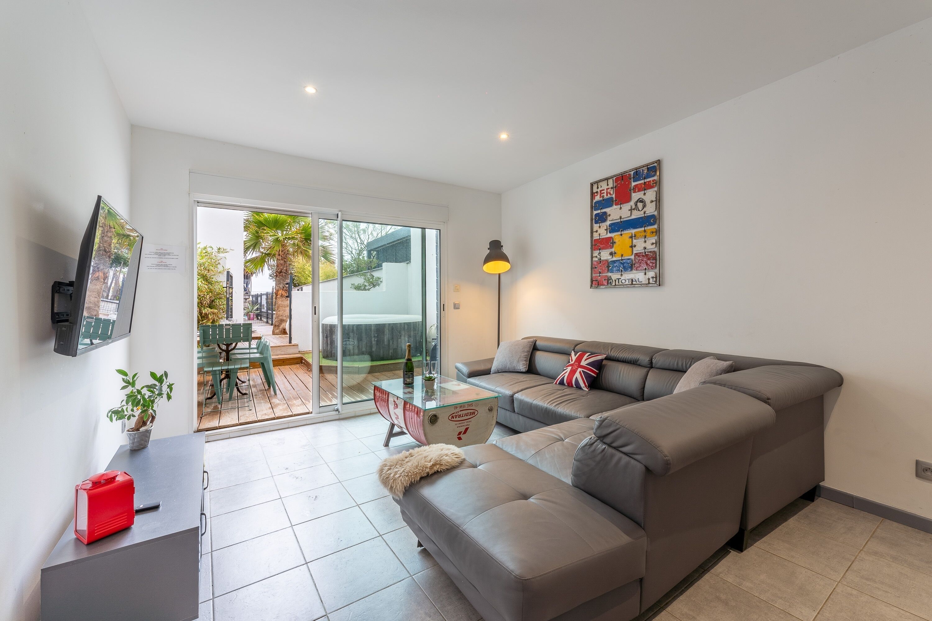 Una sala moderna con un sofá seccional gris, una mesa de centro de vidrio, un televisor de pantalla plana montado en la pared y un ventanal con vistas al exterior.