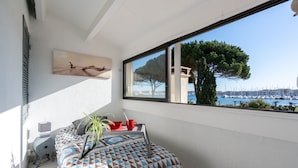 A balcony with a bed, a laptop, a red mug, and a bowl. There is a window with a view of a tree and a marina. - VILLA HESTIA - Vue sur la Marina (Agde)