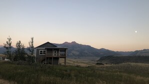 A two-story house with a balcony, surrounded by a grassy field and mountains in the background.