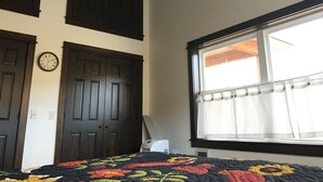 A bedroom with a large window, a bed with a colorful quilt, a ceiling fan, and a wall clock.